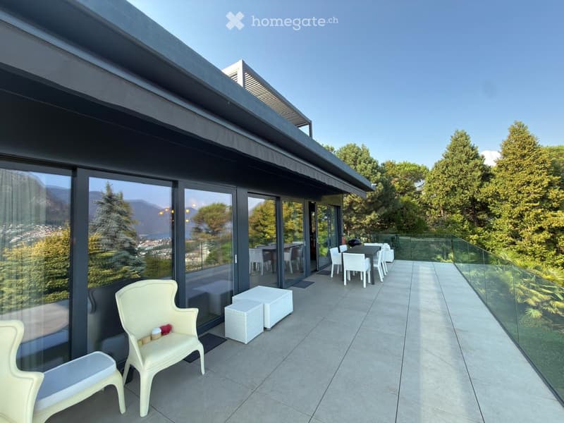 Modern balcony with large glass sliding doors, white plastic chairs and stools, a small table, and a scenic mountain view.