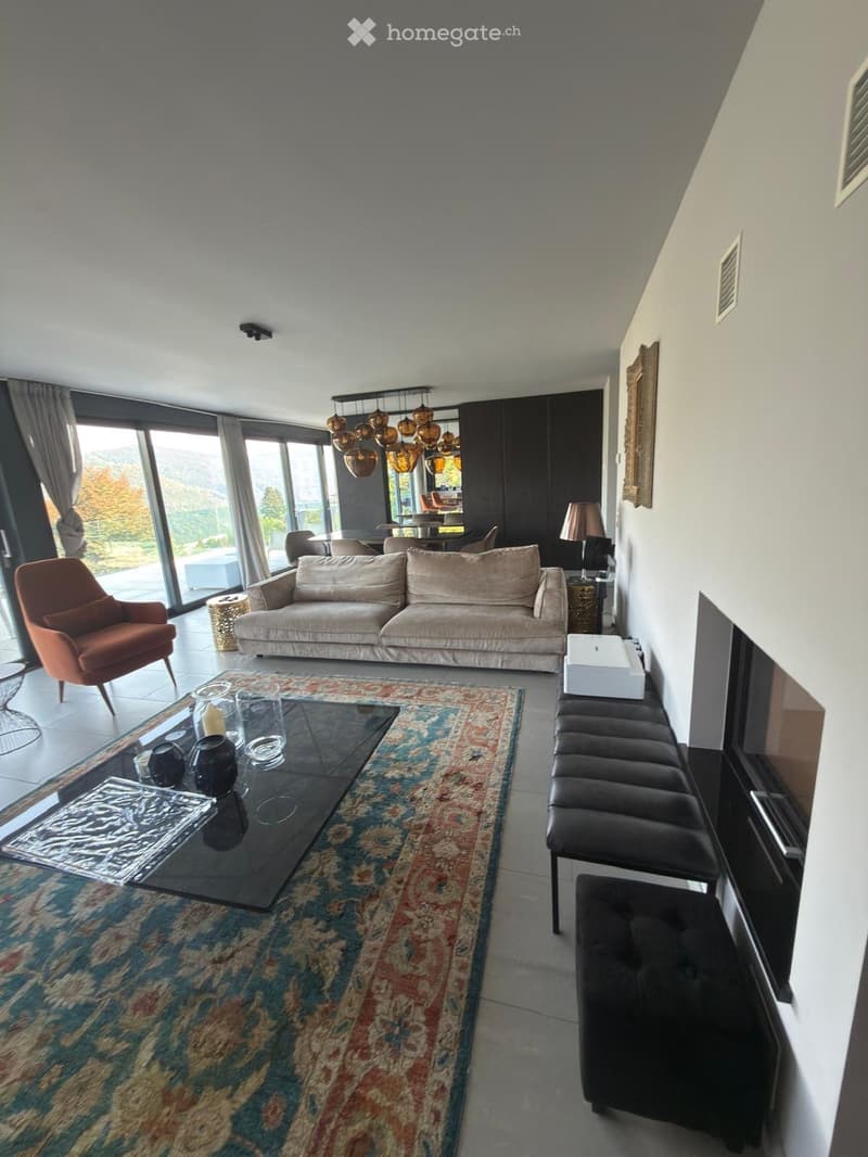 Modern living room with large windows offering a scenic view, featuring a beige sofa, glass coffee table, orange armchair, bench seating, and decorative items.