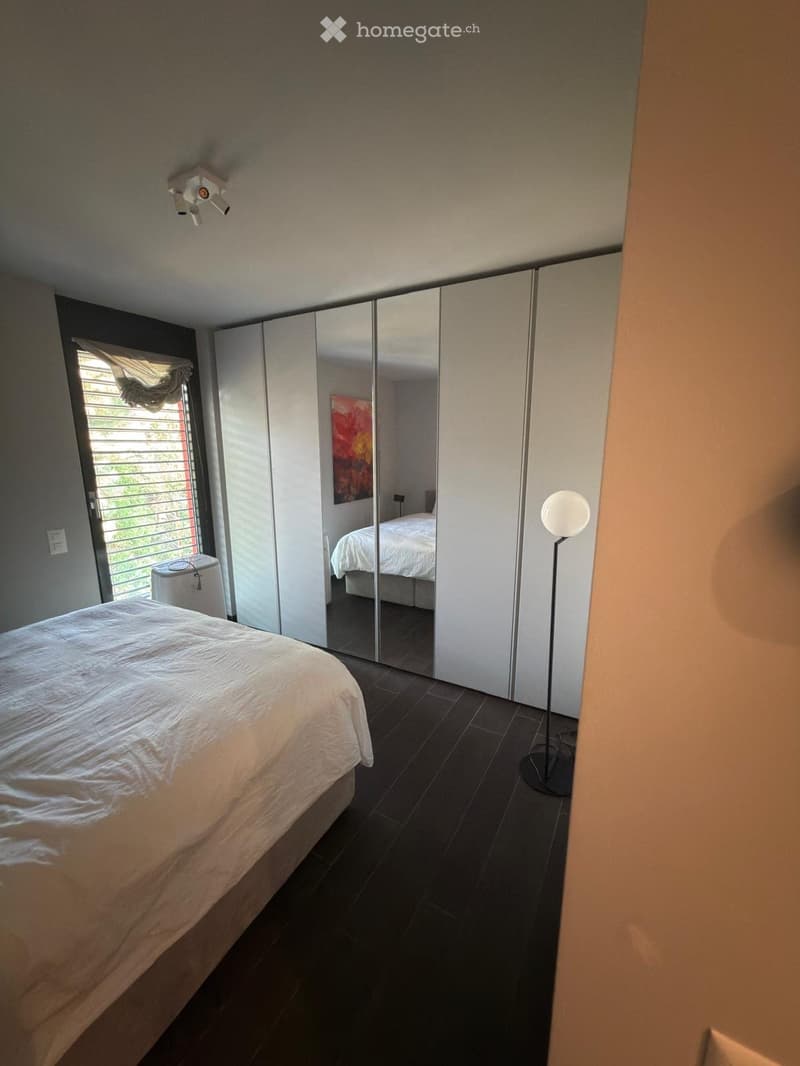 Modern bedroom with a large bed, mirrored wardrobe, floor lamp, and a painting on the wall. The room features dark tiled flooring and large windows with blinds.