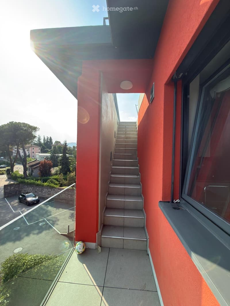 A narrow outdoor corridor with red walls, concrete steps leading upwards, a glass railing on one side, and a view of a parking area and greenery.
