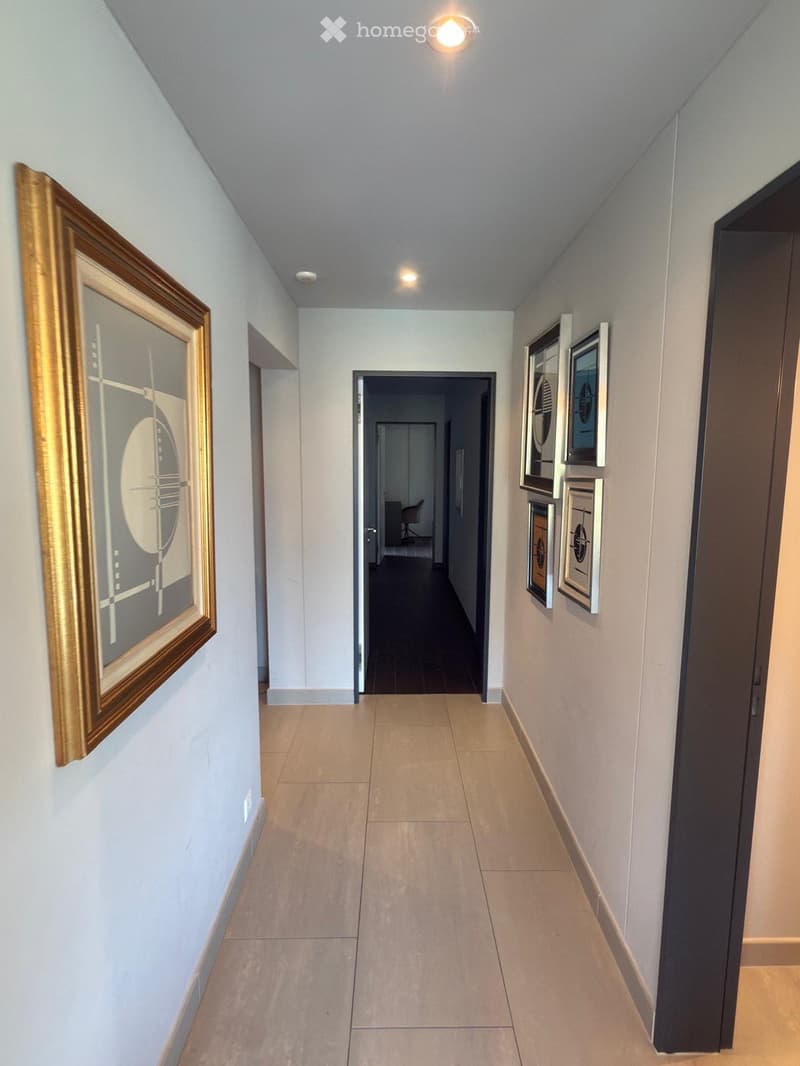 A clean, well-lit hallway with beige tiled flooring, light blue walls, and several framed artworks. The hallway features recessed lighting and leads to an open doorway at the end.