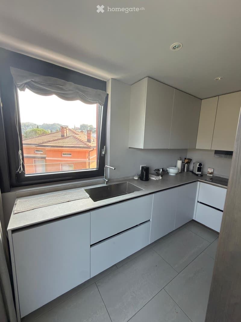 Modern kitchen with white cabinets, stainless steel sink, and various appliances on the counter, with a large window overlooking a tiled roof area.