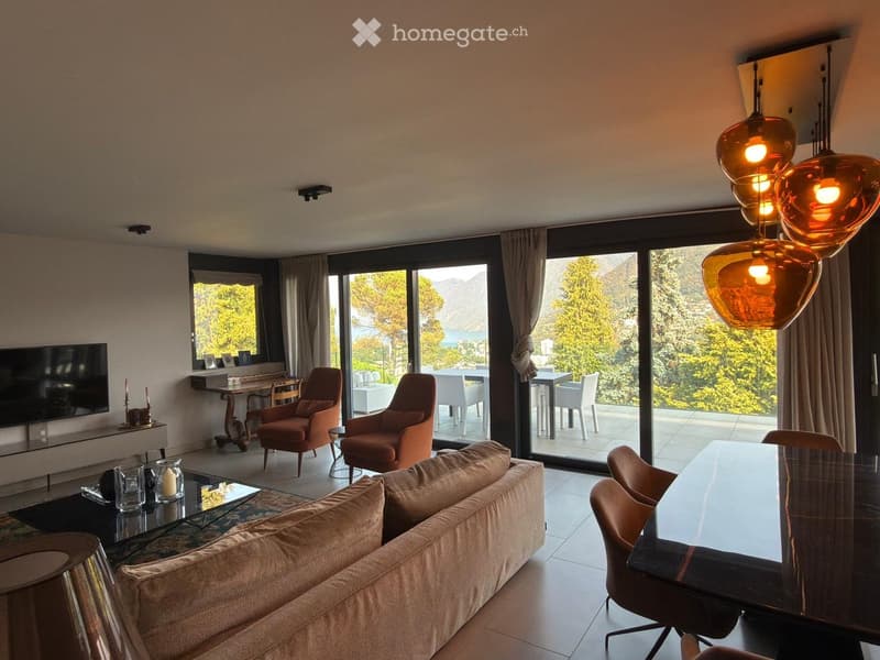 Modern living room with large windows offering a scenic view, featuring a beige sofa, glass coffee table, two armchairs, a dining table with chairs, and a television mounted on the wall.