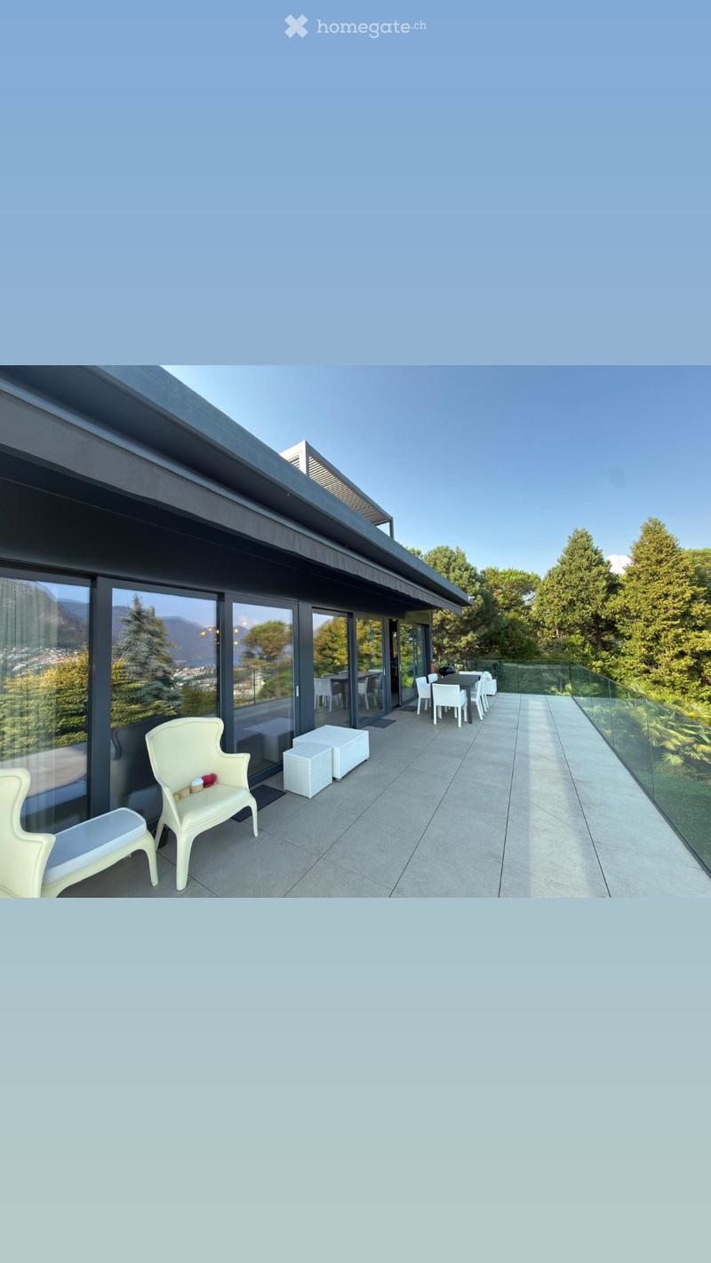 Modern balcony with large glass windows, white chairs, stools, and a small table, overlooking a scenic view of trees and mountains.