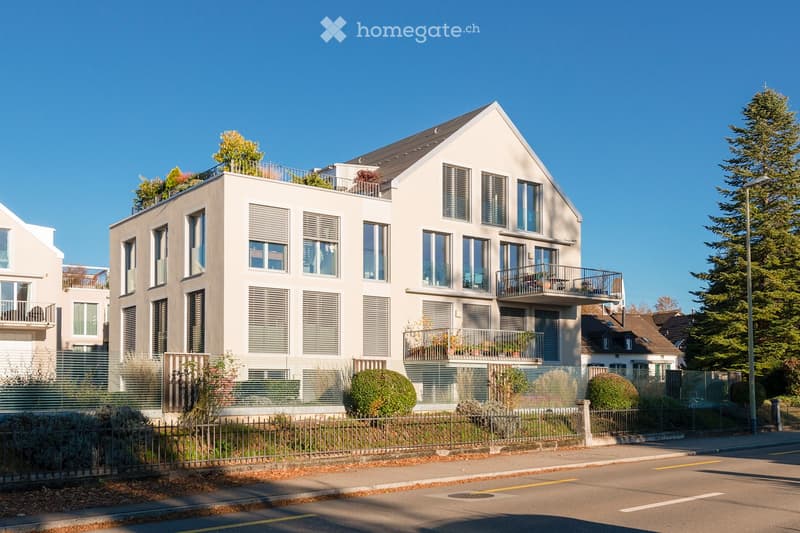 Modern multi-story residential building with large windows, balconies, and a rooftop terrace, surrounded by greenery and a fence.