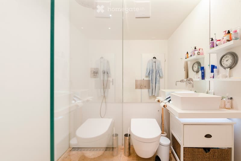 Modern bathroom with white fixtures, two sinks, a bathtub, and a glass-enclosed shower area. Shelves hold toiletries and a clock is mounted on the wall.
