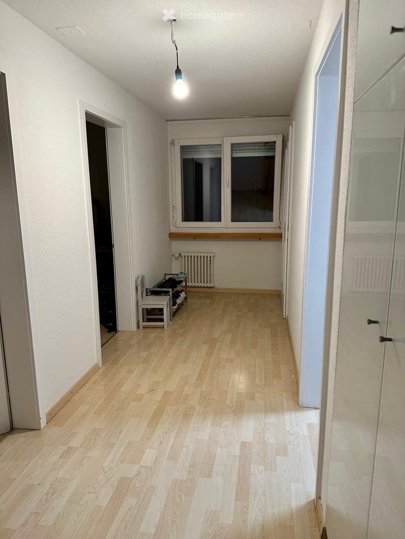 Empty hallway with light wooden flooring, white walls, and a single hanging light fixture. A small bench and shoe rack are visible on the left side, and a window with blinds is at the far end.