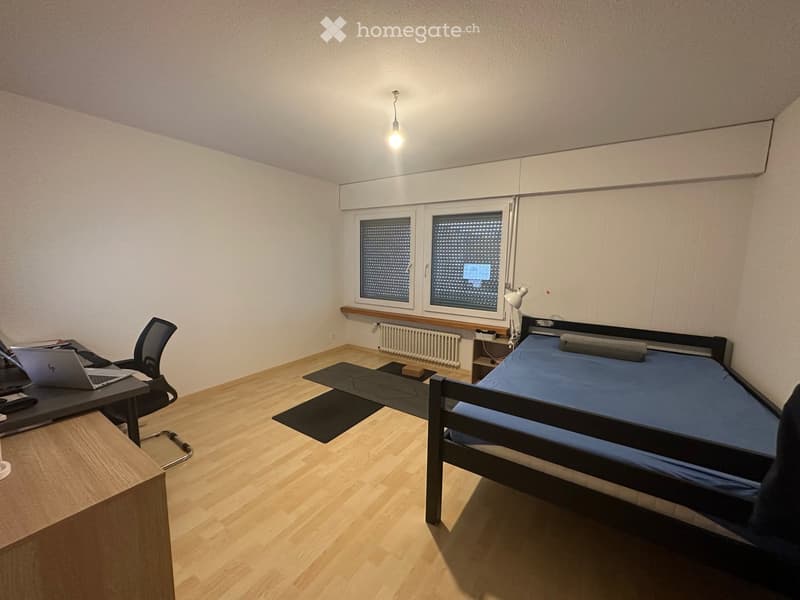 A furnished bedroom with a bed, desk, laptop, chair, and a lamp. The room features wooden flooring and large windows with blinds.