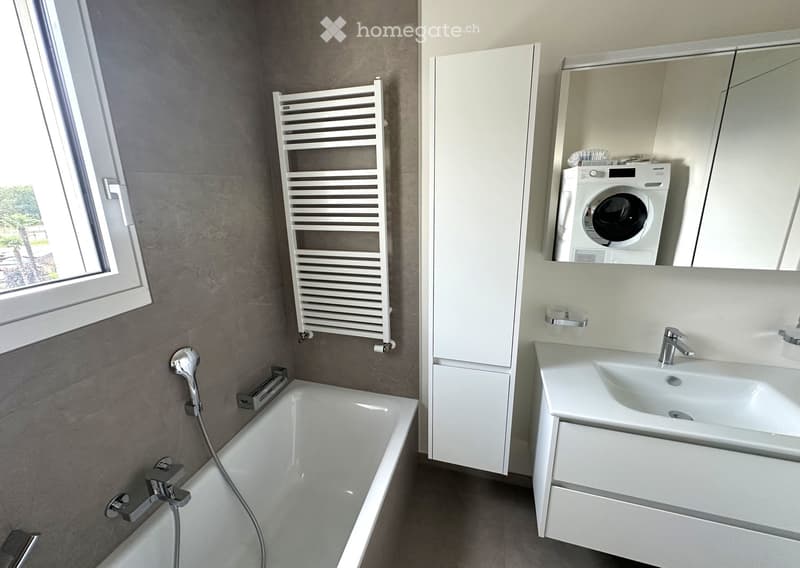 Modern bathroom with a bathtub, sink, mirror, radiator, and a washing machine in the corner.