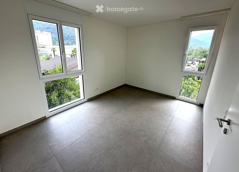Empty room with grey tiled floor, white walls, large windows offering a view of greenery and buildings, and a door with a silver handle.