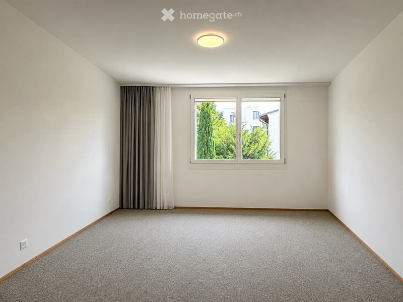 Empty room with white walls, carpeted floor, large window with curtains, and a ceiling light fixture.