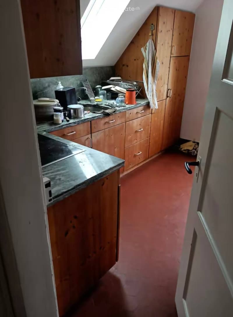 The kitchen features wooden cabinets, a marble countertop, and a sink with a faucet. Various kitchen items, including a microwave, oven, and cups, are visible on the counter. The floor is red, and there is a window with a skylight above the sink.
