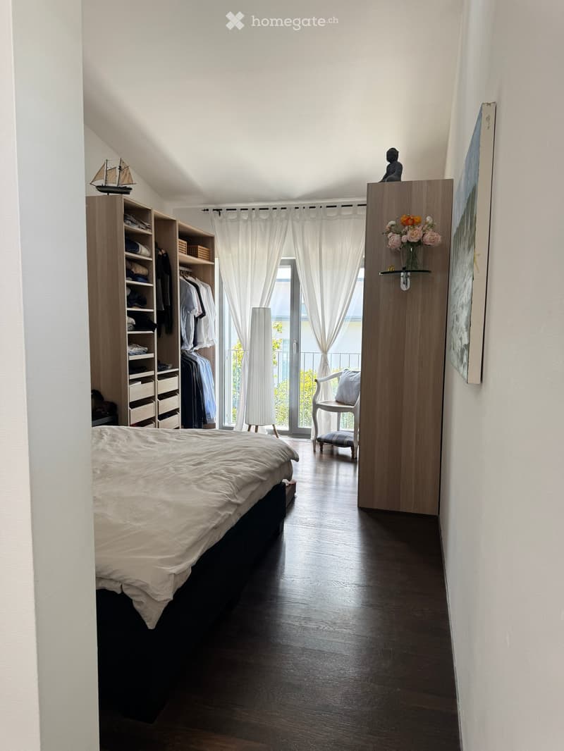 Bright bedroom with wooden floor, large bed, built-in wardrobe, white curtains, and a view of a balcony.