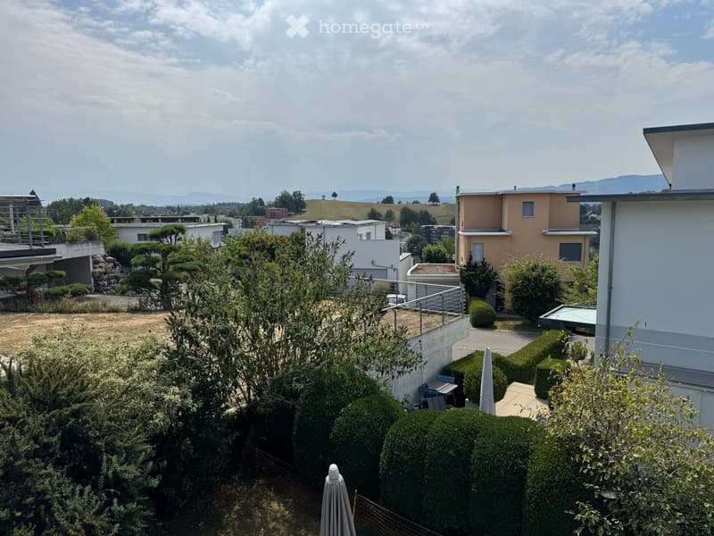 A well-maintained garden with trimmed hedges, trees, and a small patio area with outdoor furniture, overlooking a suburban neighborhood with modern houses and distant hills.