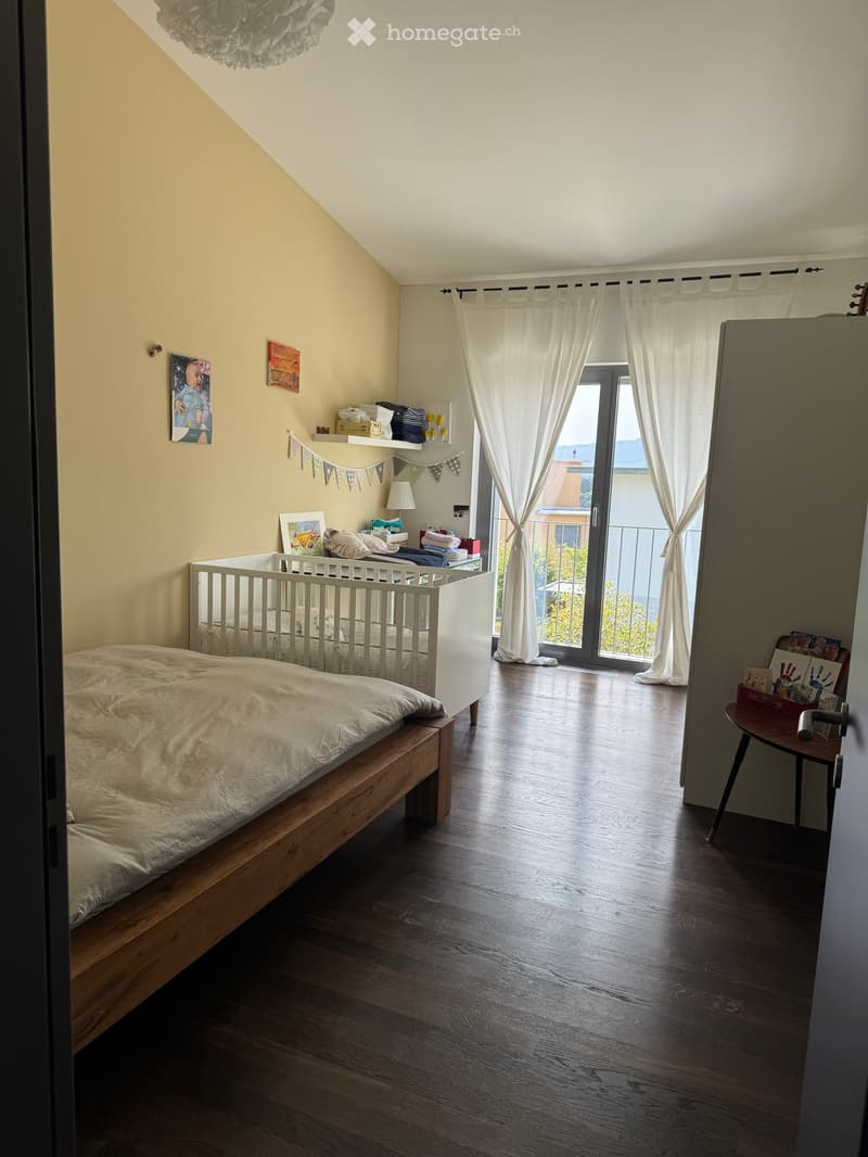 Bright bedroom with wooden flooring, large window with white curtains leading to a balcony, a double bed, a crib, a small table, and shelves with decorative items.
