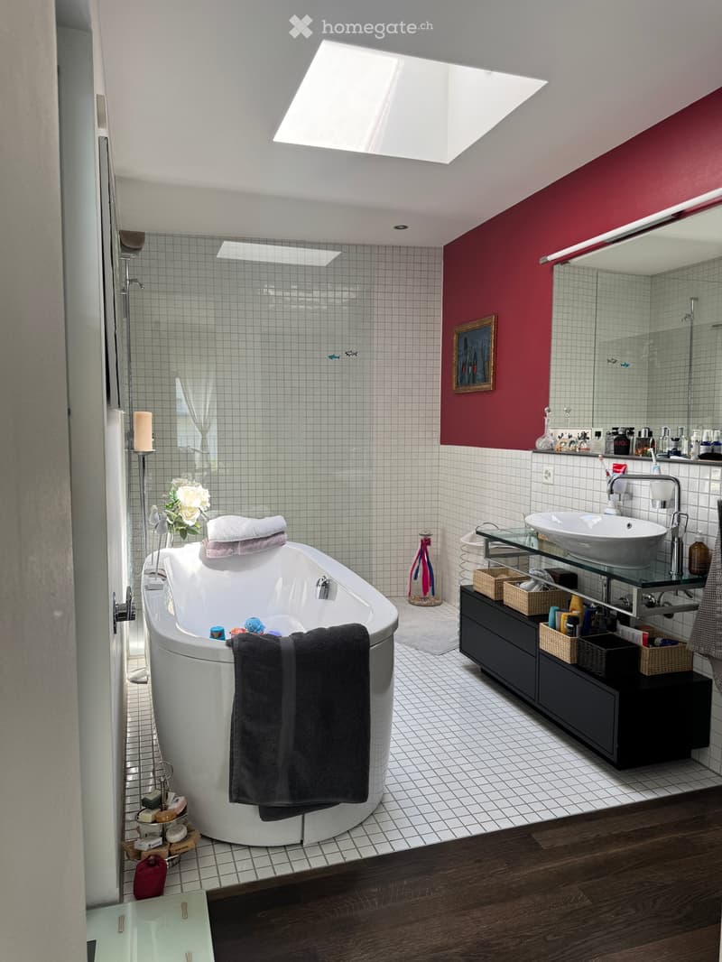 Modern bathroom with a large bathtub, sink, mirror, and various toiletries. Features a red accent wall, white tiled shower area, and a skylight window.