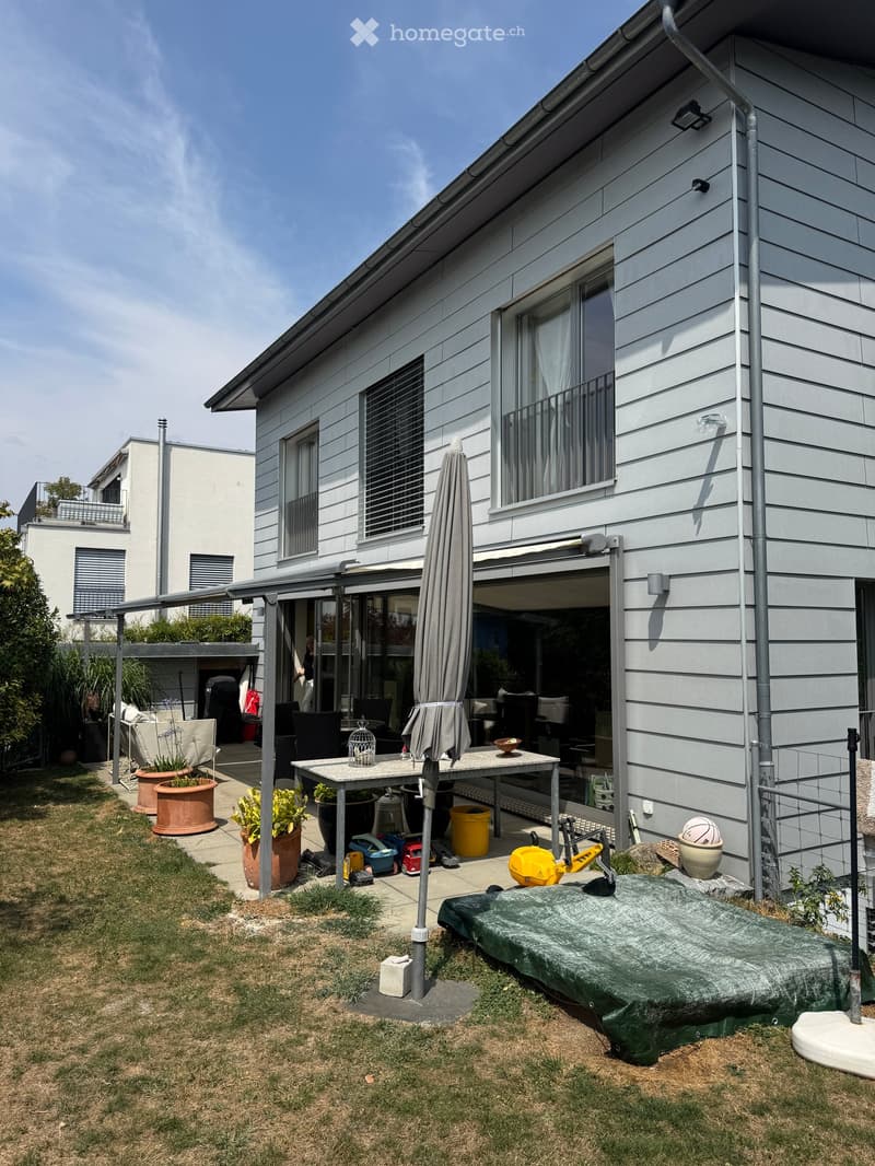 Outdoor patio area with a table, chairs, umbrella, potted plants, and a covered swimming pool. The patio is attached to a modern house with large windows and a balcony.
