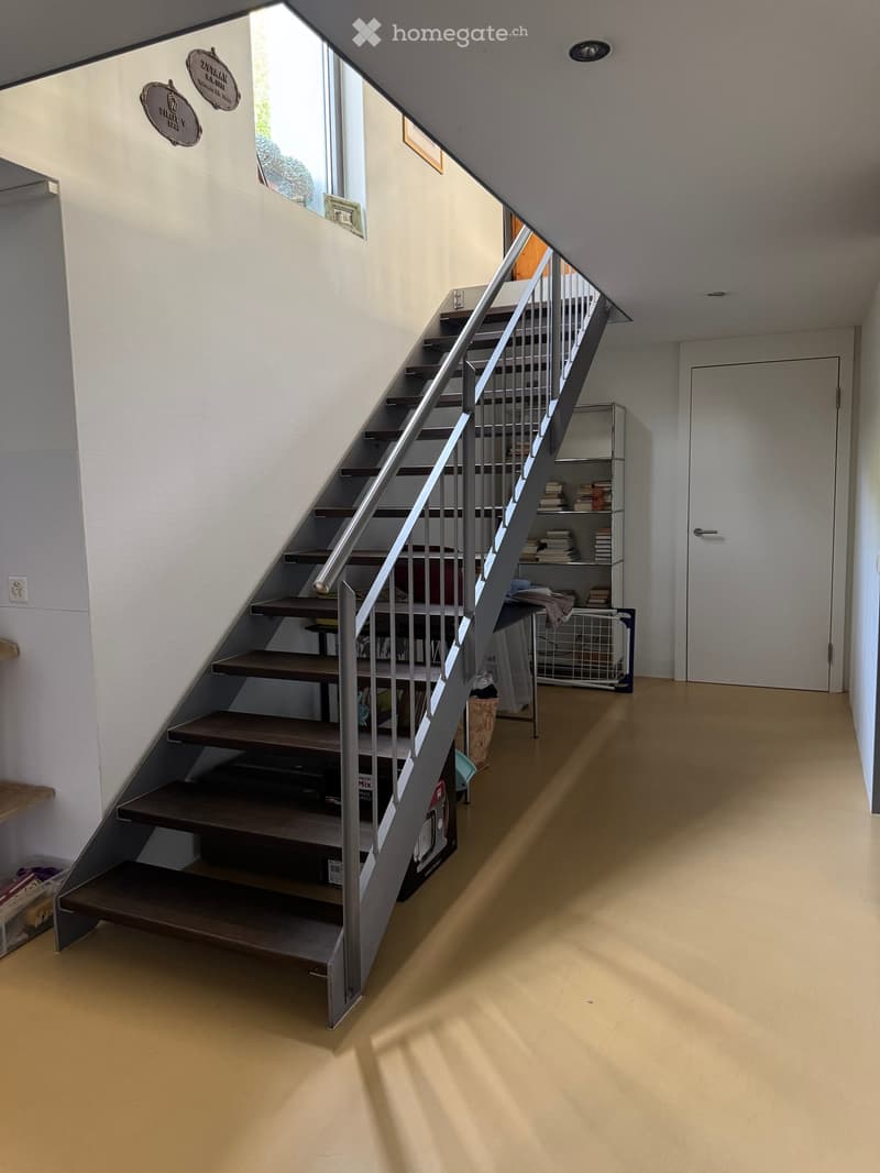 Modern hallway with a wooden staircase featuring a metal railing, a small shelf with books, and a closed door at the end.