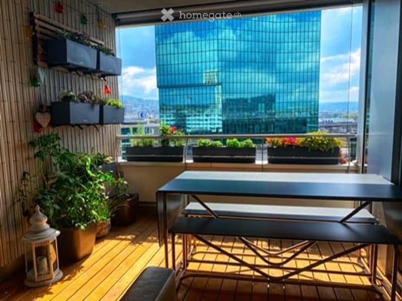 Balcony with wooden flooring, outdoor table and benches, potted plants, and a view of a modern glass building.