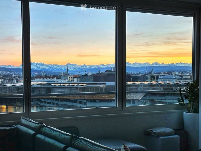 A modern balcony with large windows offering a panoramic view of a cityscape and mountains at sunset. A green sofa and a potted plant are visible.