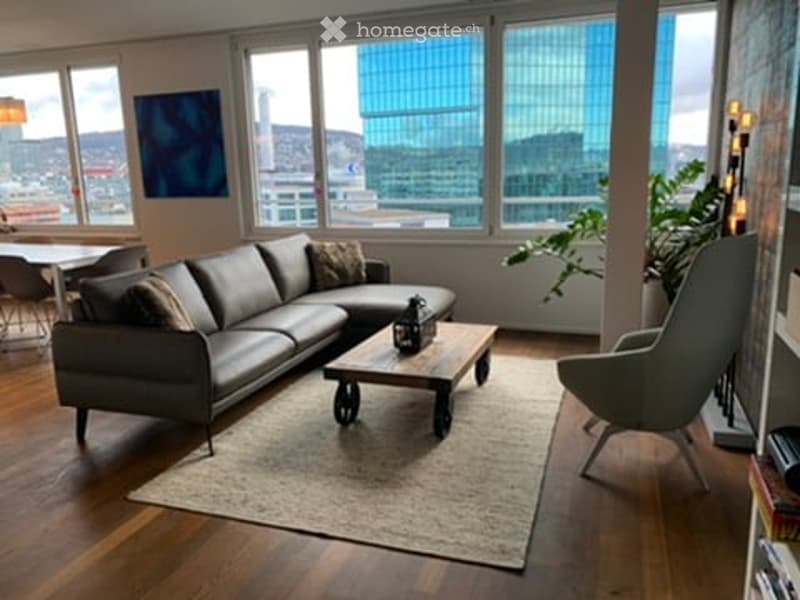 Modern living room with large windows offering a city view, featuring a grey sectional sofa, wooden coffee table, armchair, dining area with table and chairs, and decorative plant.
