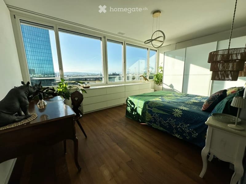 Modern bedroom with large windows offering a city view, featuring a bed with a patterned duvet, a nightstand with a lamp, a wooden table with a chair, and decorative plants.