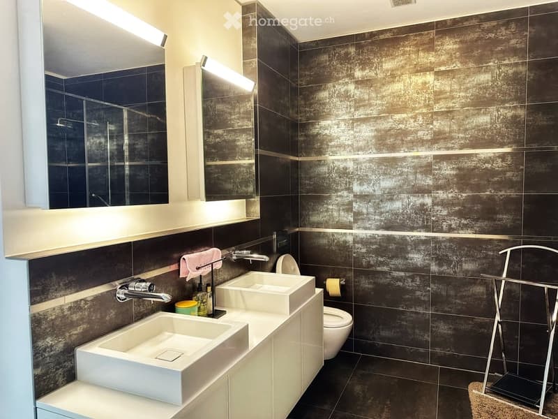 Modern bathroom with dark tiled walls, two white sinks, a toilet, a mirror, and a towel rack. The design features sleek, contemporary fixtures and ample lighting.