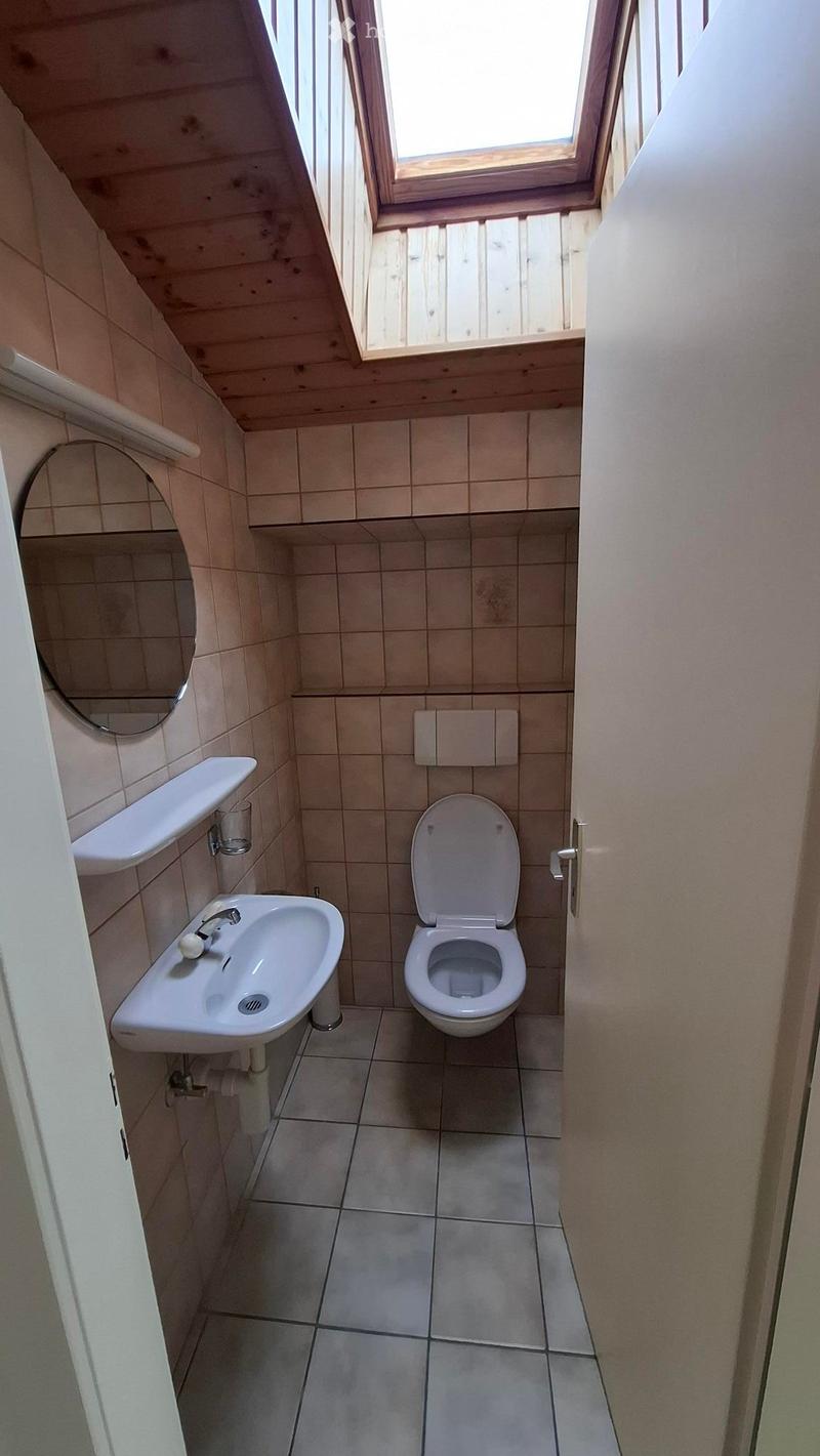 Small bathroom with a toilet, sink, mirror, and a cup on the shelf. The room has tiled walls and a skylight window.