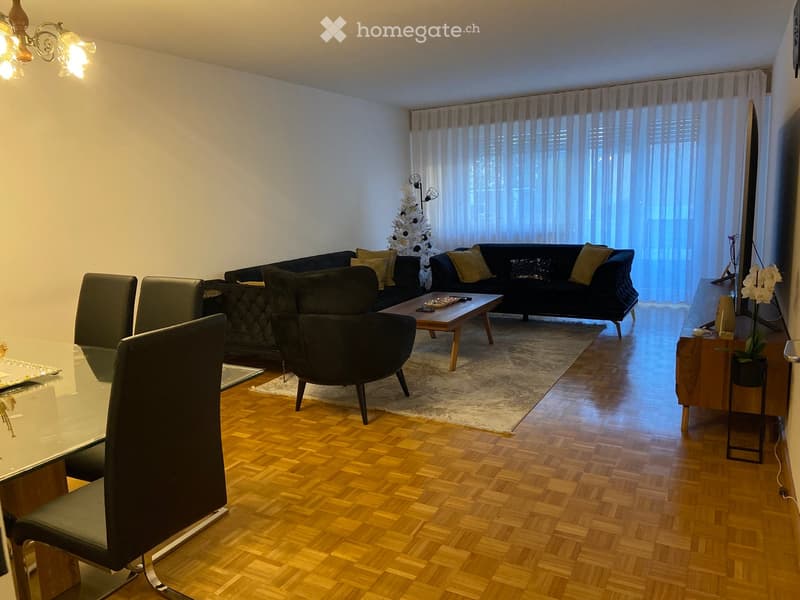 Modern living room with wooden flooring, large windows with sheer curtains, a dark-colored sofa, a coffee table, armchairs, a dining table with chairs, and a television on a wooden stand.