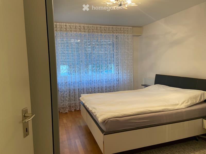 A modern bedroom with a large bed, white bedding, a nightstand, and sheer curtains on a window. The floor is wooden, and the walls are painted white.