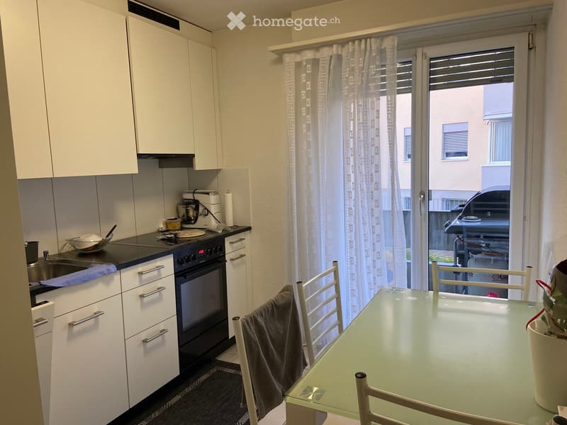 Modern kitchen with white cabinets, stainless steel appliances, a glass dining table, and chairs. A window with a curtain leads to a balcony.