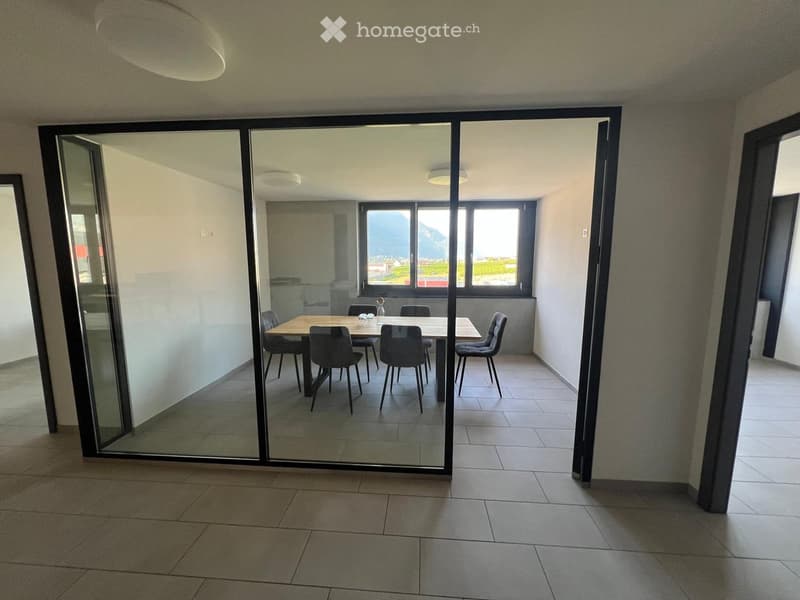 Modern dining area with a wooden table and six chairs, large windows with a scenic view, and a glass sliding door leading to another room.