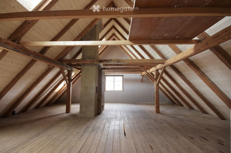 An empty attic space with exposed wooden beams and a slanted wooden ceiling. The floor is made of wide wooden planks, and there is a single window allowing natural light to enter.