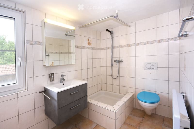 Modern bathroom with white tiled walls, a grey vanity with a sink, a bathtub, a toilet with a blue seat, a showerhead, and a mirror with built-in lighting.