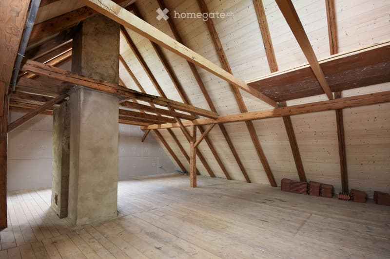 An empty attic space with wooden beams and exposed wooden floorboards, featuring concrete pillars and a partially finished wall.