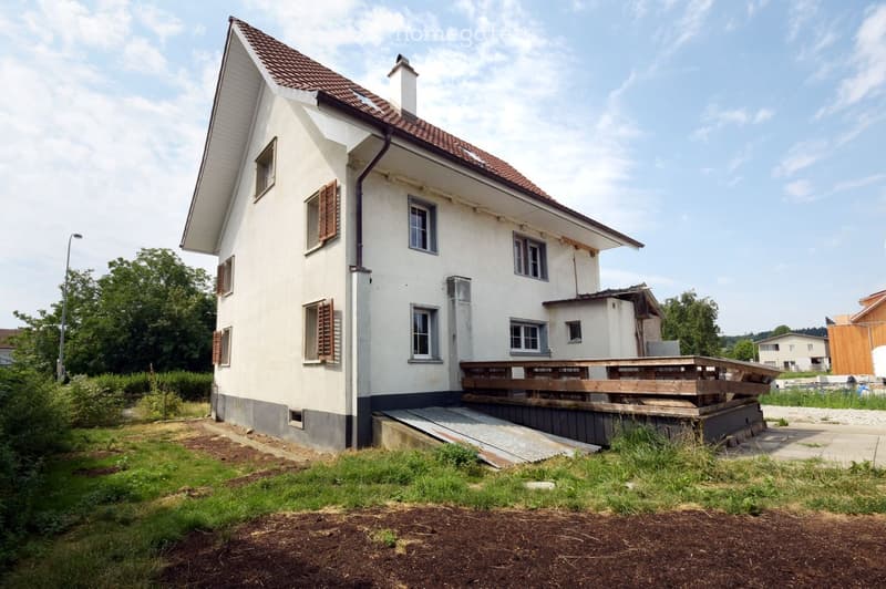 Two-story house with white exterior, brown shutters, and a wooden deck on the raised ground floor.