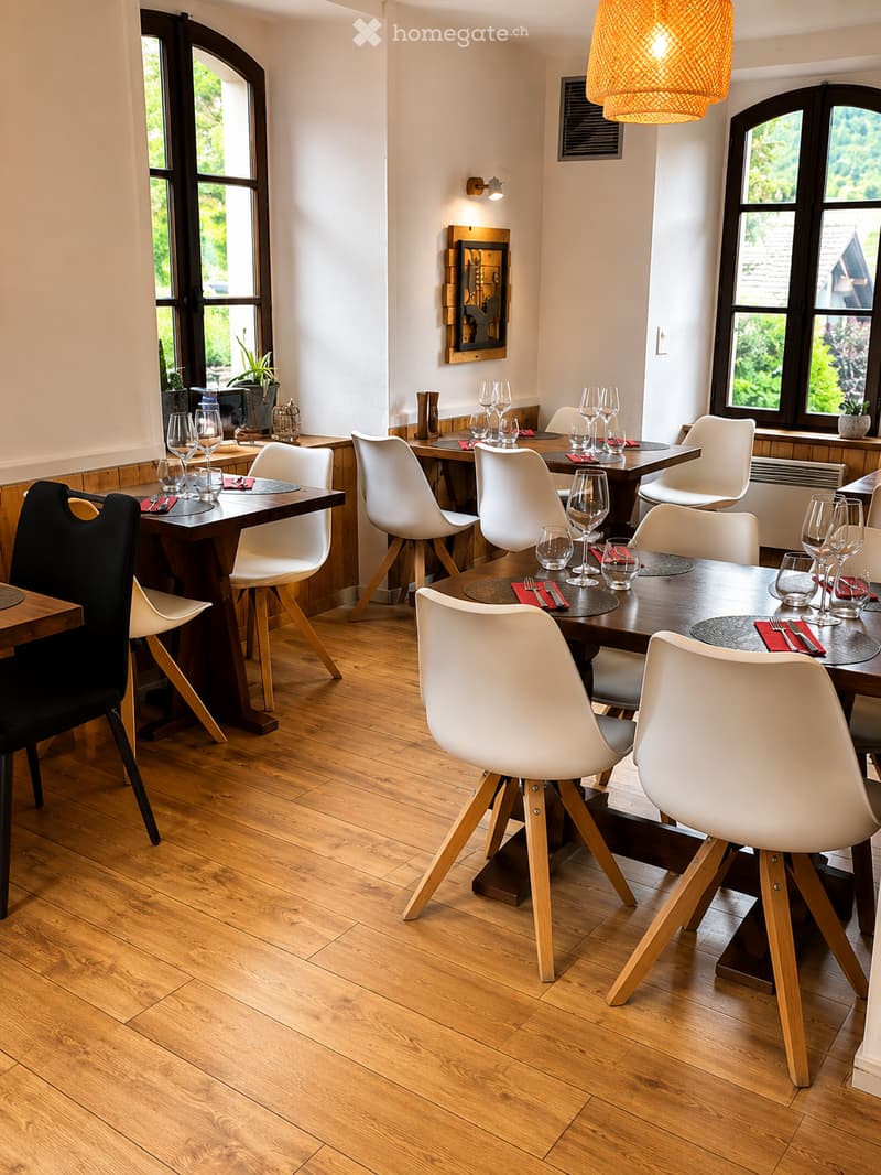 Modern dining area with wooden floors, large windows, multiple tables and chairs, wine glasses, cutlery, and decorative lighting.