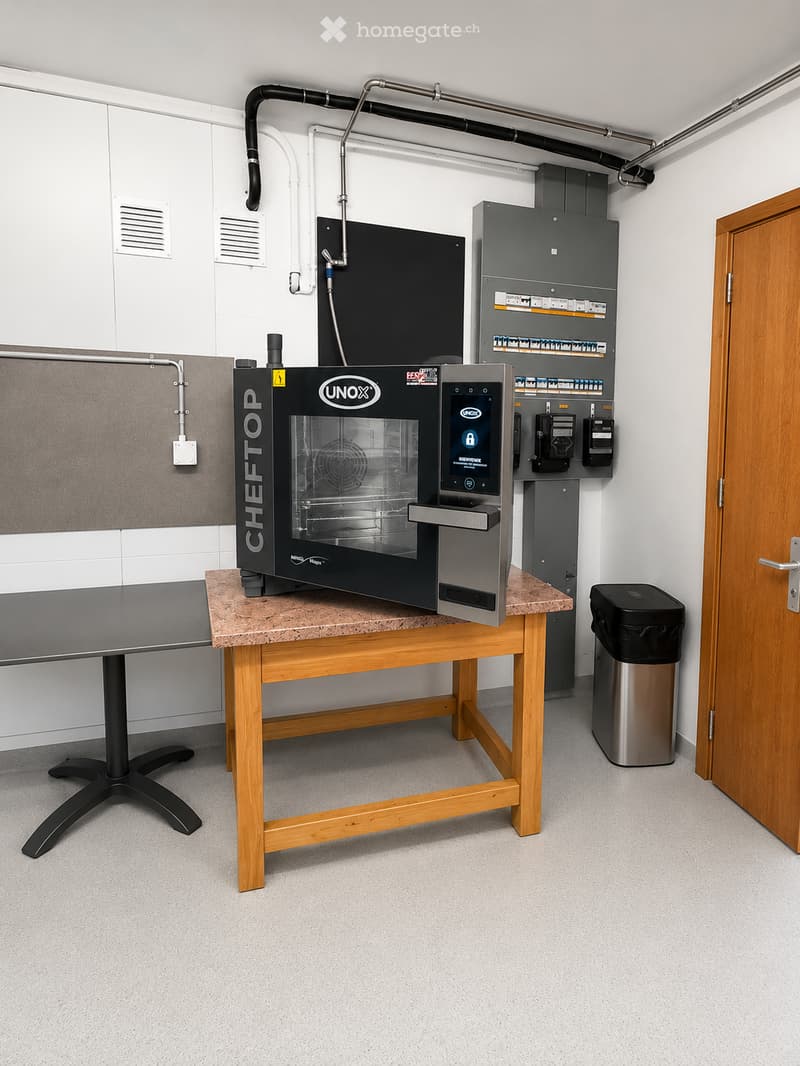 A small, well-lit room with a wooden table holding a UNOX ChefTop oven, an electrical panel, and a trash bin. The room has white walls, a grey carpeted floor, and a wooden door.