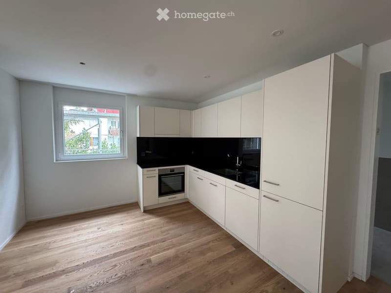 Modern kitchen with white cabinets, black countertops, and a wooden floor. Features an oven, sink, and a window with blinds.