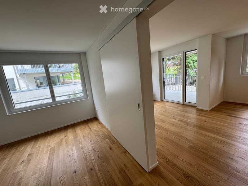 Empty room with wooden flooring, large windows, and sliding glass doors leading to a balcony.