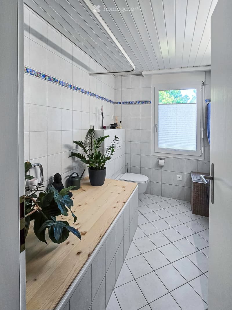 Modern bathroom with white tiles, wooden sink, toilet, and a potted plant on the sink counter.
