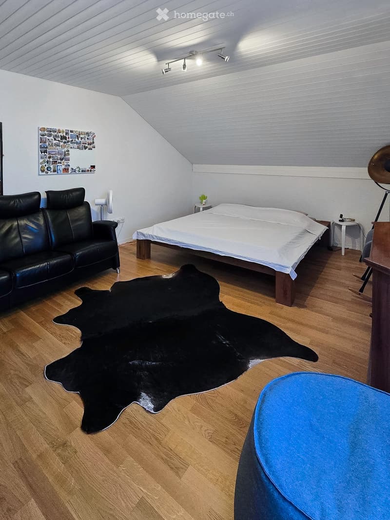A modern bedroom with a large bed, a black leather sofa, a cowhide rug, a small table with a plant, and a fan. The room features wooden flooring and white walls.