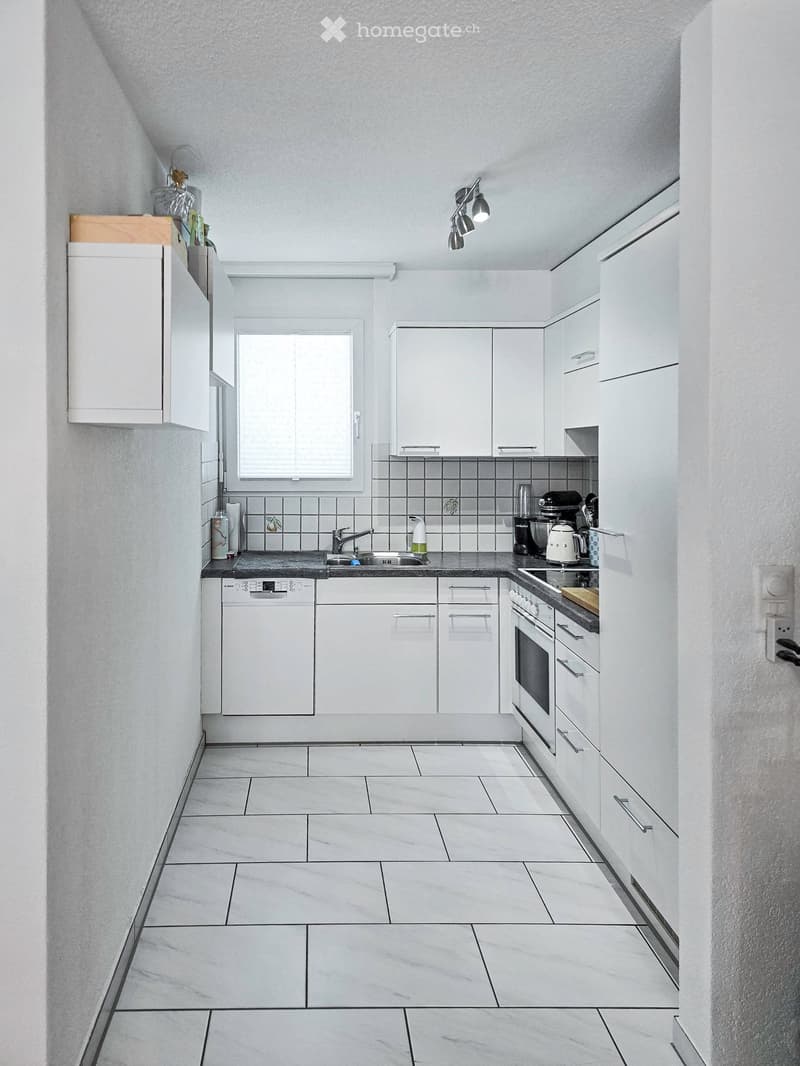 Modern kitchen with white cabinets, tiled backsplash, and stainless steel appliances including a sink, oven, and dishwasher. The space features a window above the sink and a countertop with various appliances.