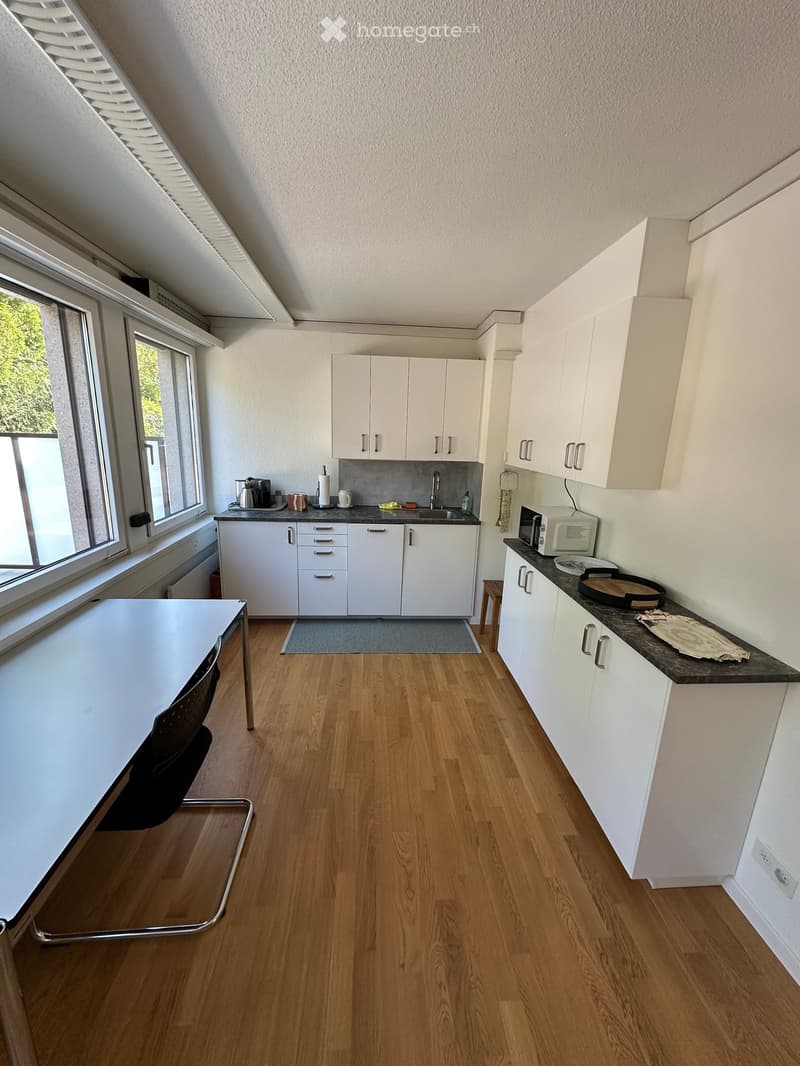Modern kitchen with white cabinets, wooden flooring, and a large window. Includes a table, chair, microwave, and various kitchen appliances.