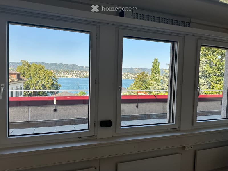 Empty room with large windows offering a scenic view of a lake and surrounding landscape. The room features white walls, a radiator, and a window sill.