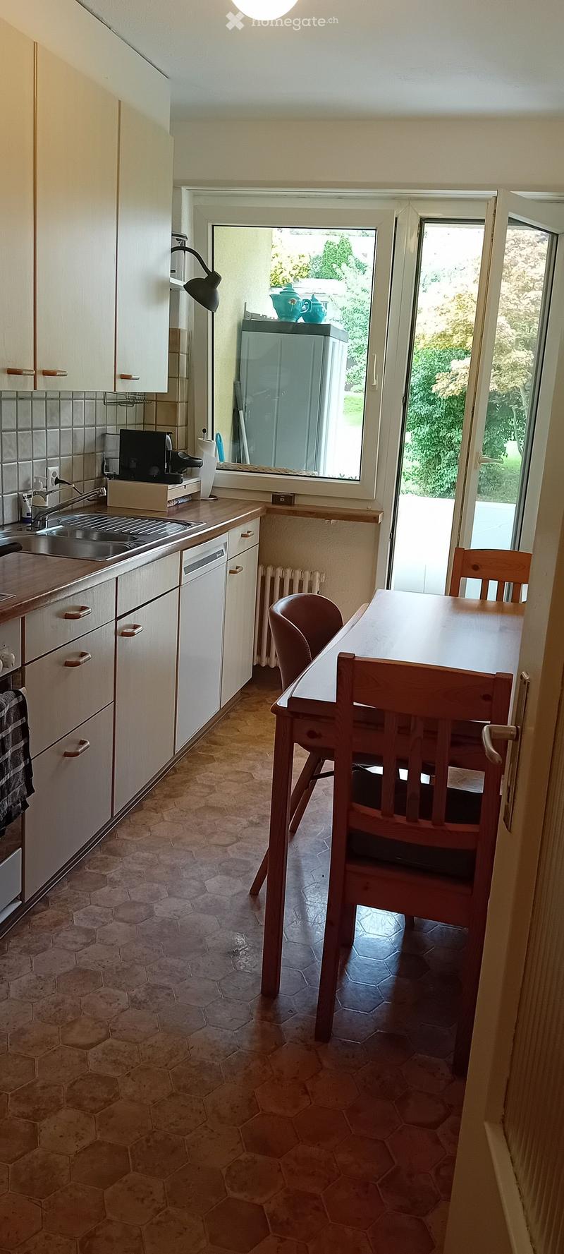 A compact kitchen with white cabinets, a sink, a dishwasher, a refrigerator, a small dining table with chairs, and a lamp on the wall.