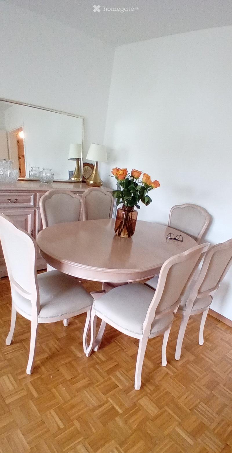 Dining room with a round wooden table surrounded by six white upholstered chairs, a vase with orange flowers, and a mirror on the wall with lamps and decorative items.