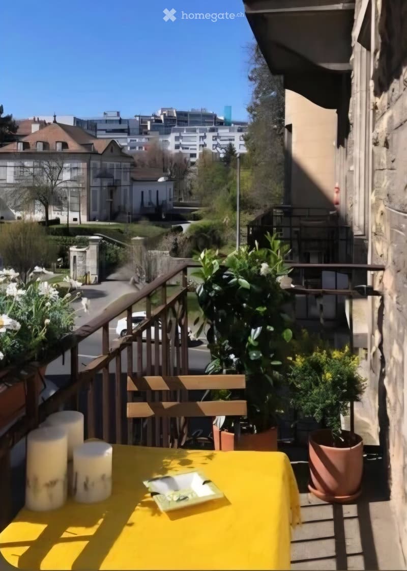 Balcony with a yellow table, chairs, candles, a plate, and potted plants, overlooking a cityscape with buildings and greenery.