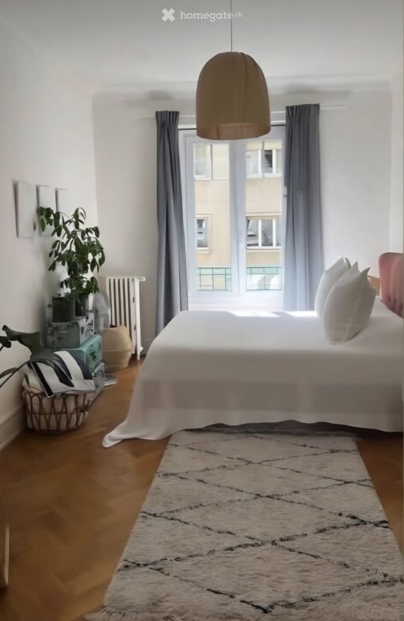 Modern bedroom with a large bed, pendant light, large window with curtains, plant, radiator, and a basket on the floor.