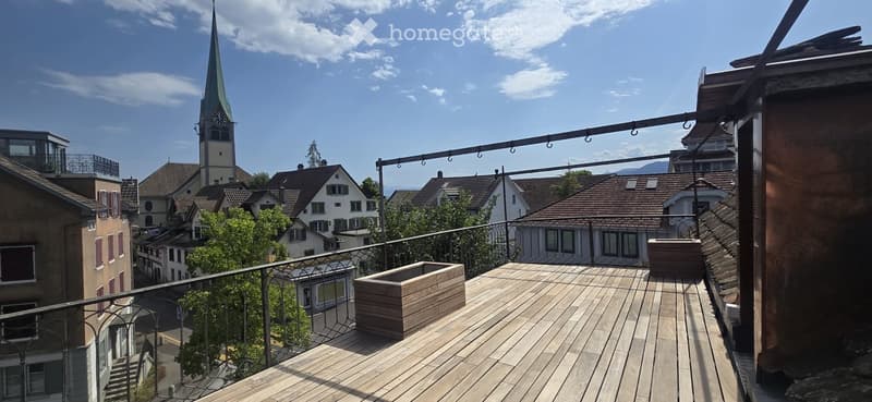 Wooden balcony with metal railing, overlooking a town with a church tower and surrounding houses.